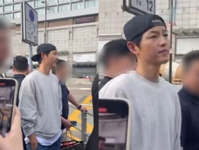 Song Joong Ki wearing a gray sweatshirt and black cap greeting fans outdoors